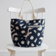 A navy and natural colored circular printed fabric tote with zipper pocket and webbed handles sitting on a white wooden chair in front of a white wall
