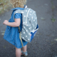 A small child wearing a blue dress and a blue and white backpack.