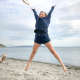 A woman in a navy blue sweatshirt and blue shorts with a red pocket detail jumping in a starfish shape on a beach.