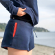 Side view of a woman in a navy blue sweatshirt and blue shorts with a red pocket detail standing on a beach. The photo is cropped to show the woman from the shoulder down to the mid-thigh.