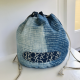A round, blue drawstring bag sitting on a while table. The bag has an oval patch of darker blue appliqué on the side.