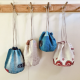 Four drawstring bags hanging from wooden pegs against a white wall. Two of the bags are blue, one is a natural linen colour, and the last is white with red embroidery. The drawstrings are different lengths so the bags each hang at a different hight against the wall.