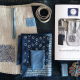 A tie on pocket of linen embellished with blue appliqué laying flat on a dark surface with a paper copy of a pattern, some folded scraps of blue fabrics, blue twill tape and a card of blue embroidery floss.