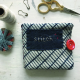 A needle book made of navy and white fabric with an appliqué rectangle of navy blue and the word &amp;quot;stitch&amp;quot; on the front and a red button closure laying on a white painted surface with a pair of embroidery scissors, a couple of locking stitch markers, a spool of string, and a card of blue embroidery floss.