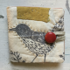 A needle book laying on a white painted surface. It is made of cream coloured fabric with a grey birds pattern. The bird has been outlined in pale yellow embroidery thread. There is a appliqué rectangle of yellow fabric with matching embroidery at the top and a large red button closure on the tail of the bird.