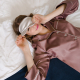 Photo of woman lying in bed wearing a sleep mask and lifting it up so one eye is visible, smiling at the camera.
